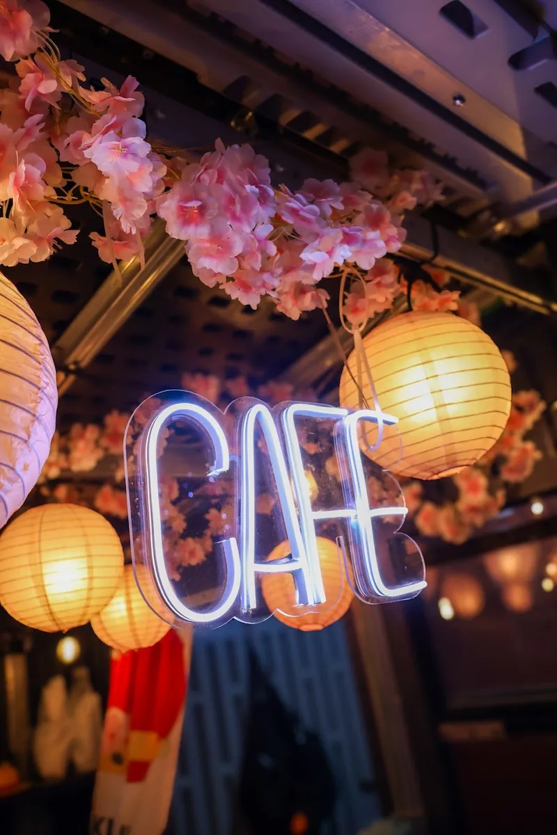 Letrero de neón con la palabra CAFÉ entre flores de cerezo y lámparas de papel encendidas en Hanami Gaviotas Sur.