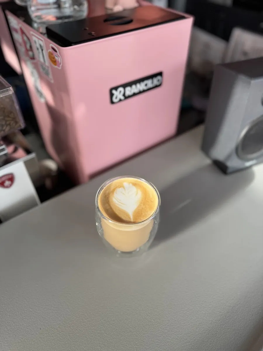 Café con leche y arte latte en vaso de vidrio, frente a la máquina de espresso rosa.